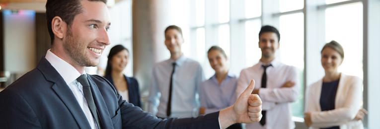 A man in a suit gives a thumbs-up in the foreground, while five people stand in the background smiling.